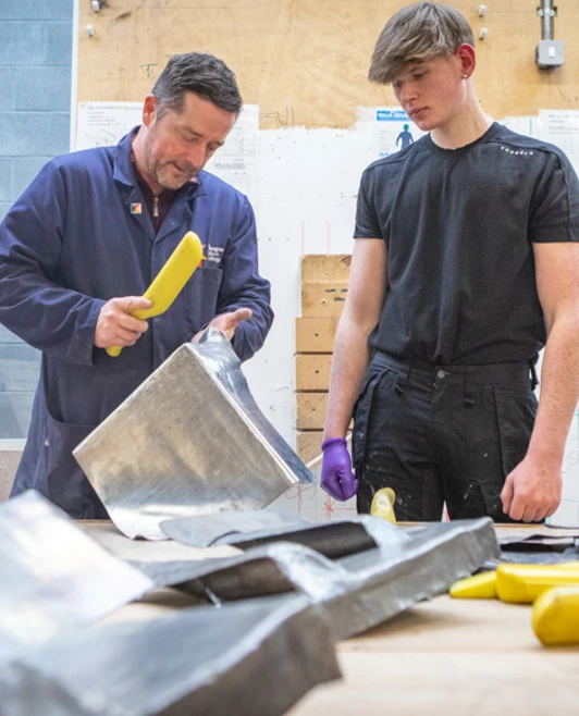 A plumbing lecturer demonstrating leadwork techniques to a student, shaping sheet metal with specialized tools in a workshop A plumbing lecturer demonstrating leadwork techniques to a student, shaping sheet metal with specialized tools in a workshop